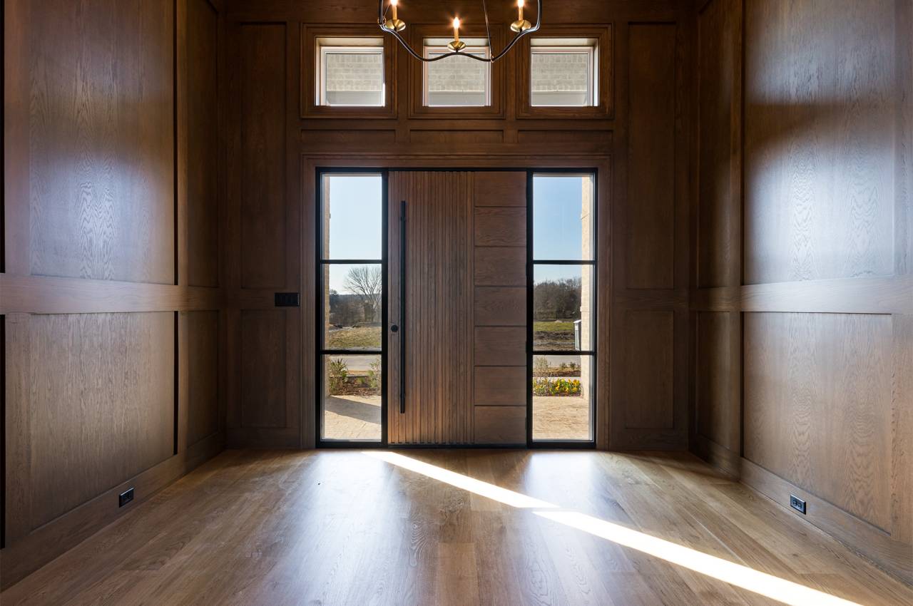 Light and bright foyer entrance with wood door and flooring, a designer chandelier, and surrounded in white oak paneling.